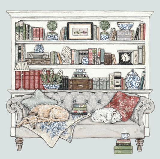 A greetings card featuring 2 dogs asleep on a button backed sofa infront of a bookcase full of old books, blue and white china and collectibles. A steaming cup of tea on a pile of books in the foreground.