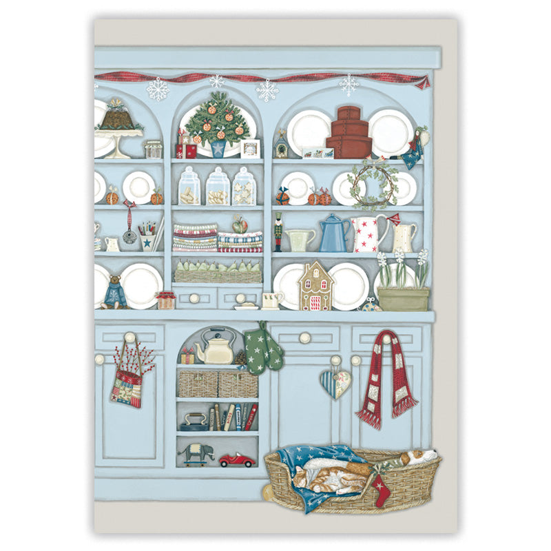 A  Christmas card featuring a whimsical illustration of a light blue dresser filled with holiday decorations, plates, jars, and small ornaments, with a sleeping dog and cat in a basket at the bottom right.