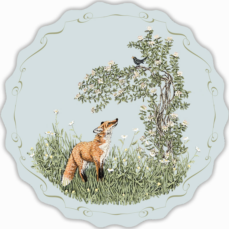 Circular postcard depicting a fox standing in long grass and wildflowers, looking up at a blackbird sat on a honeysuckle tree.
